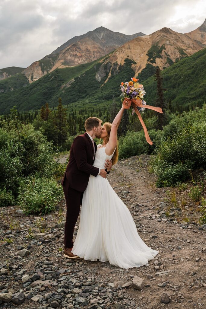 Bride raises bouquet into the air while kissing groom at the base of the mountains