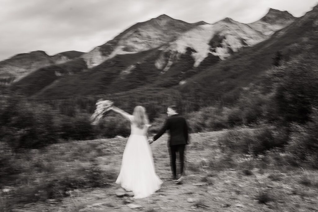 Bride and groom hold hands while running through the mountain trail in Alaska