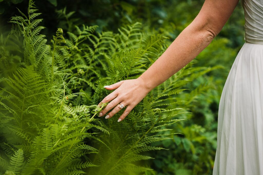 Bride runs hand along the ferns while walking through a mountain trail