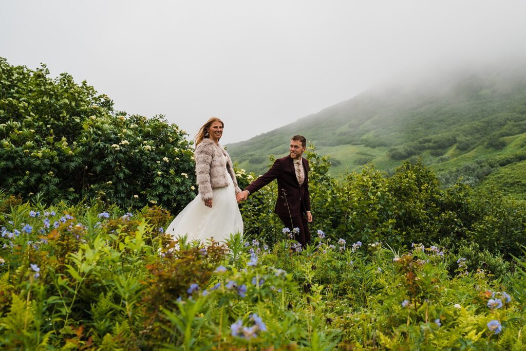Bride and groom hold hands while walking through a lush green trail in Hatcher Pass, Alaska