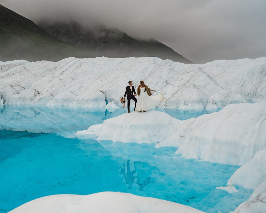 Bride and groom standing on an icy glacier surrounded by clear, blue water during their helicopter elopement in Alaska