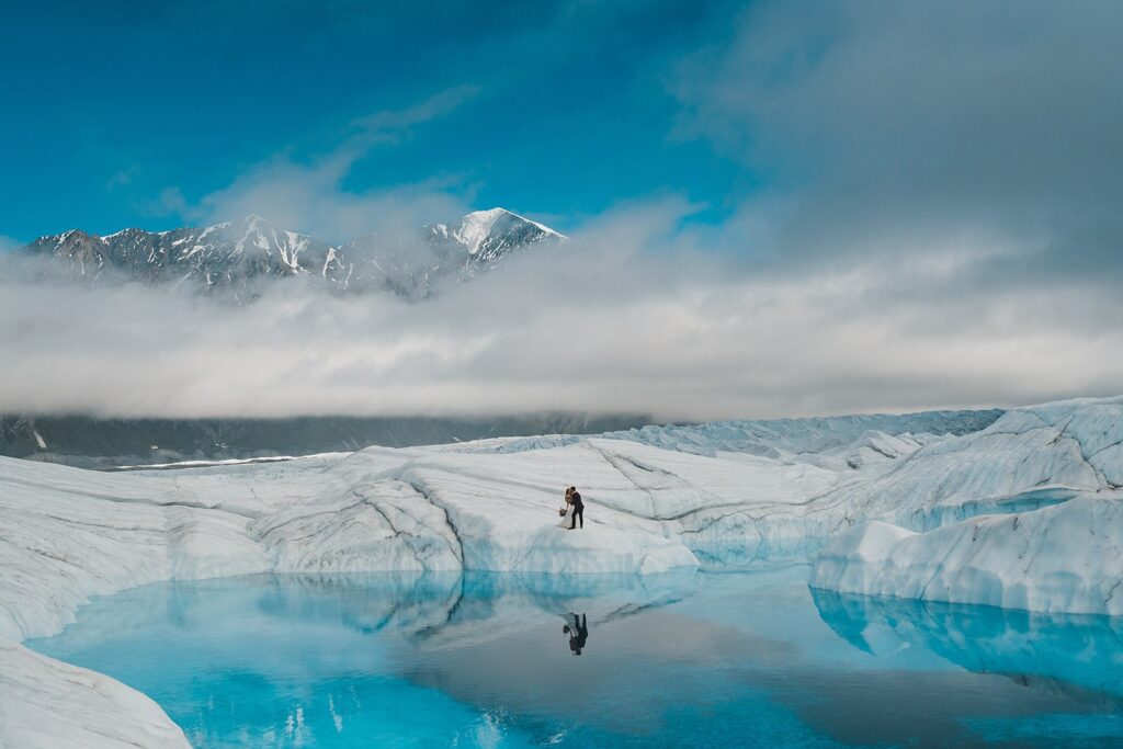 Bride and groom kiss on an icy glacier surrounded by clear, blue water during their helicopter elopement in Alaska