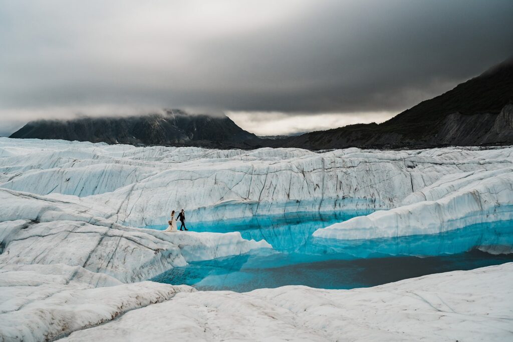 Bride and groom hold hands while walking across an icy glacier surrounded by clear, blue water at their helicopter elopement in Alaska