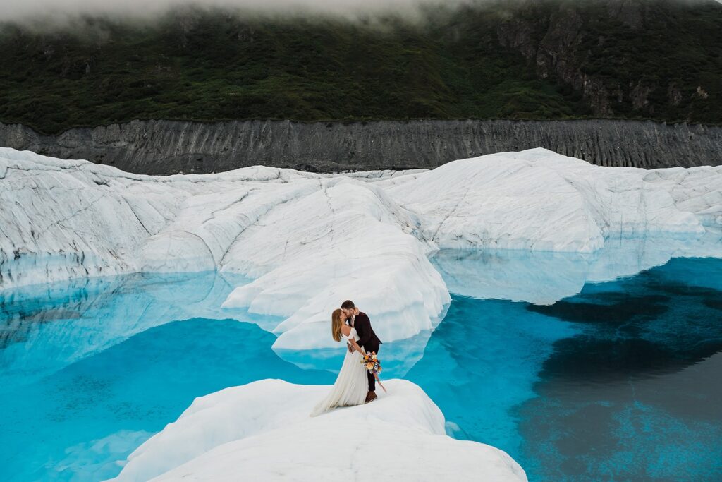 Bride and groom kiss on an icy glacier surrounded by clear blue water during their helicopter elopement in Alaska