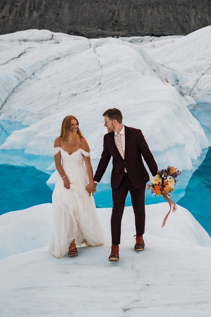 Bride and groom hold hands while walking across an icy glacier surrounded by clear, blue water during their Alaska helicopter elopement