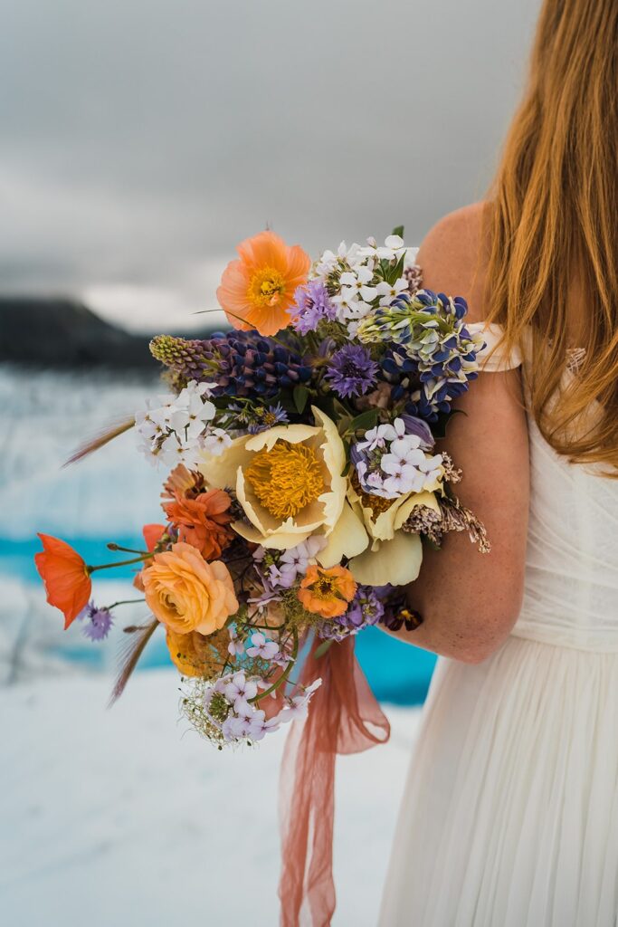 Bride holds colorful wildflower bouquet with yellow and orange flowers at her Alaska helicopter elopement