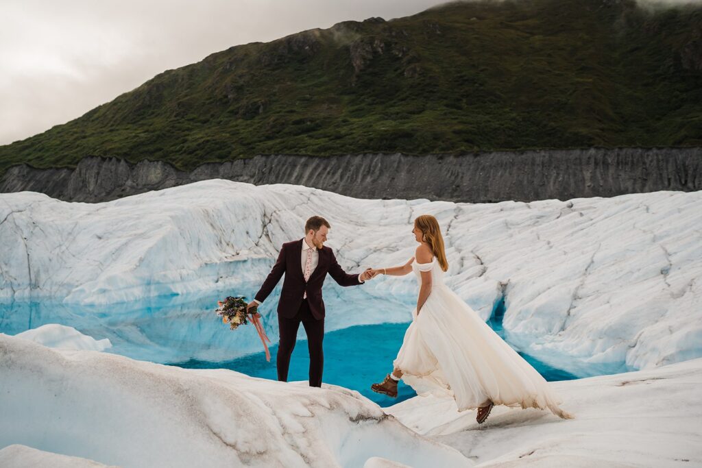 Groom helps bride over an icy glacier during their helicopter elopement in Alaska