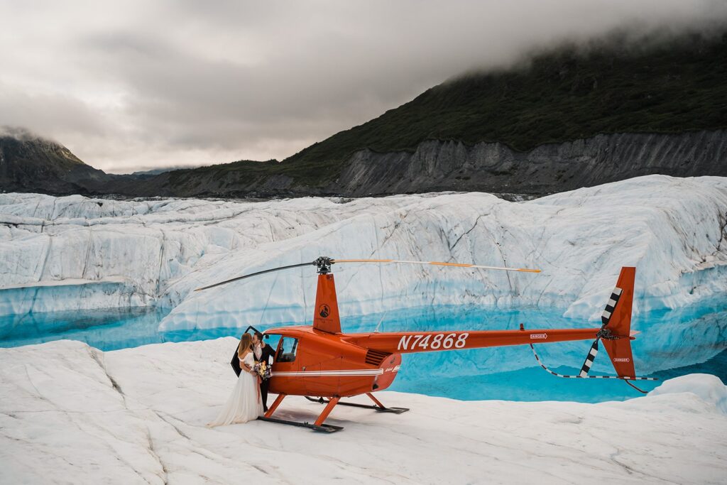 Bride and groom kiss near a red helicopter during their two-day elopement in Alaska