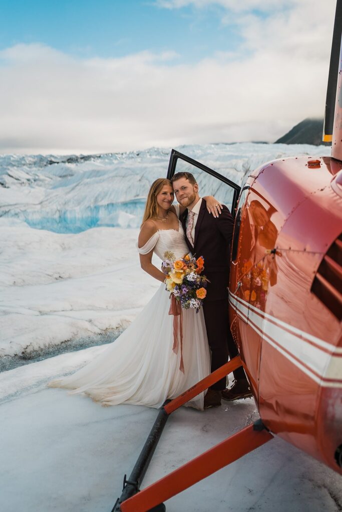 Bride and groom stand next to a red helicopter during their two-day Alaska elopement
