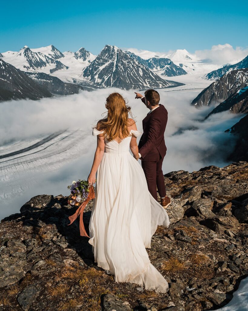 Bride and groom stand on a mountain and look out to the horizon during their helicopter elopement in Alaska