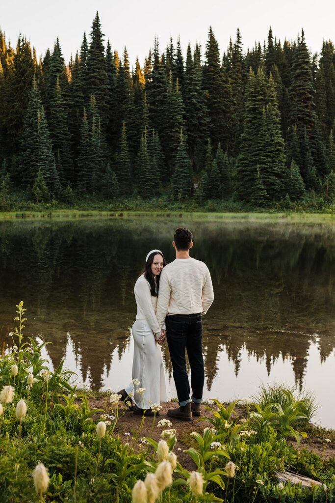 Bride and groom hold hands beside an alpine lake during their sunrise elopement