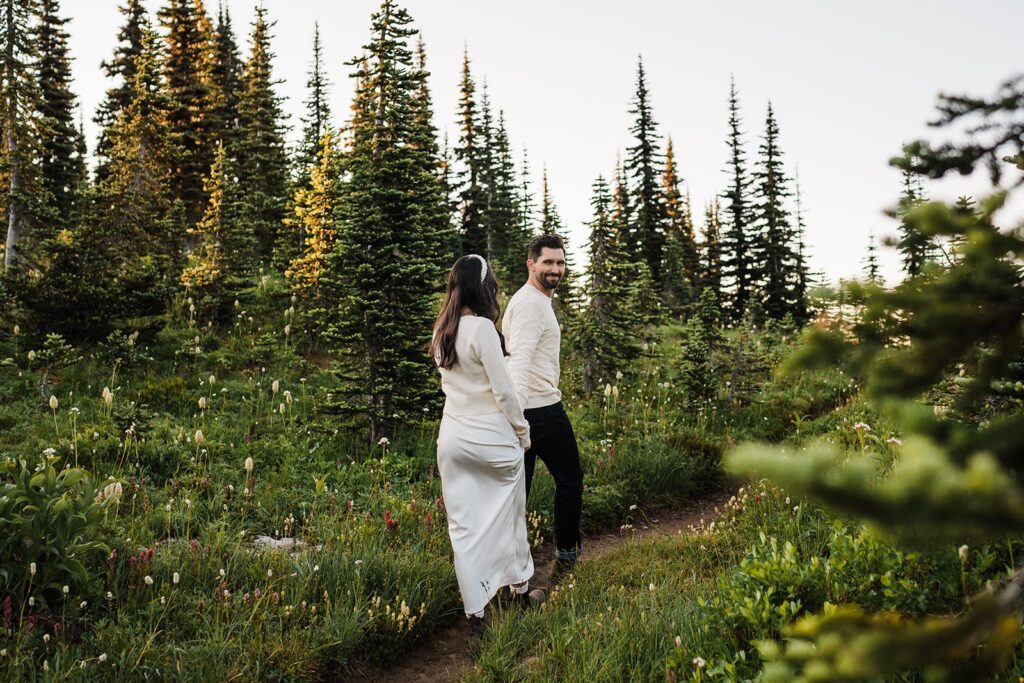Bride and groom hold hands while walking through a mountain trail surrounded by wildflowers