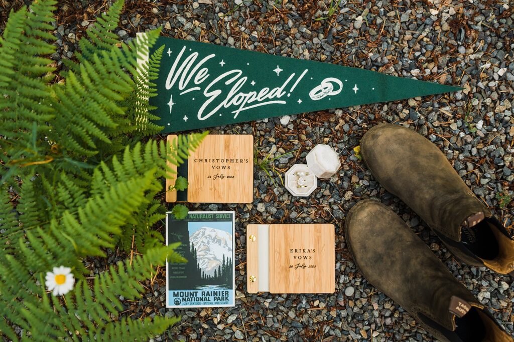 Elopement detail flatlay with gree "We Eloped" flag, wood vow books, hiking boots, and a Mt Rainier National Park postcard