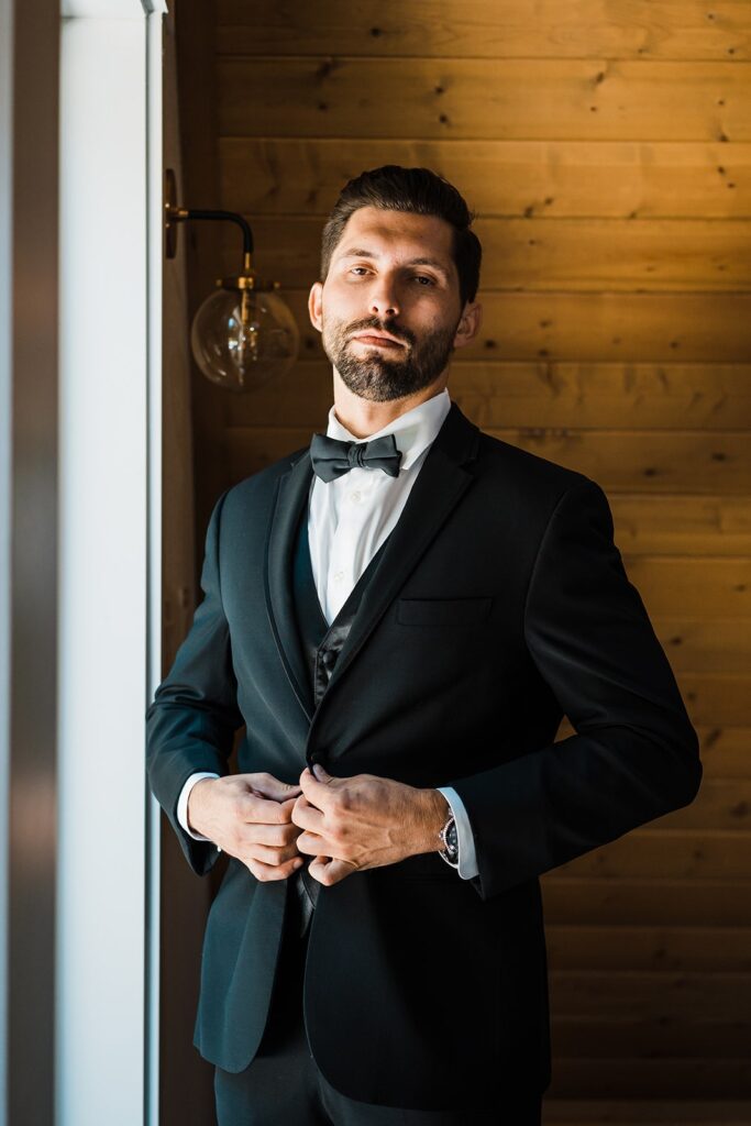 Groom buttons his black elopement suit while getting ready for his wedding first look 