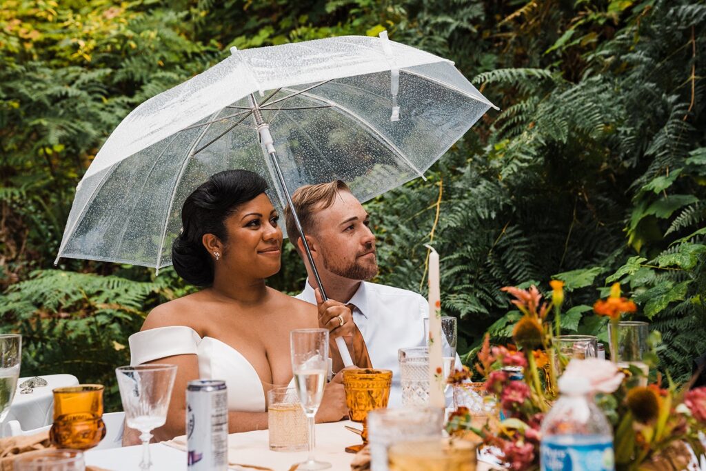 Bride and groom sit under a clear umbrella during their family elopement reception meal