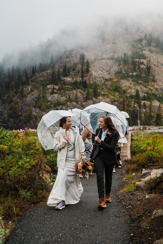 Brides hike in the North Cascades while holding clear umbrellas and wedding flowers