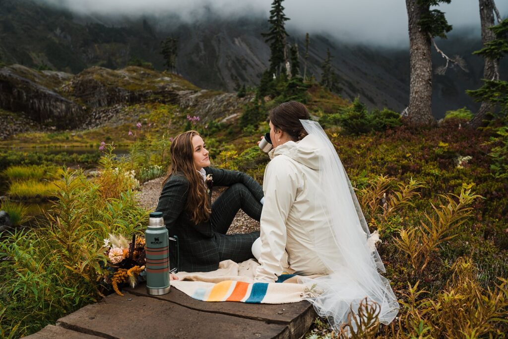 Brides sit on a blanket, sipping hot chocolate during their North Cascades wedding
