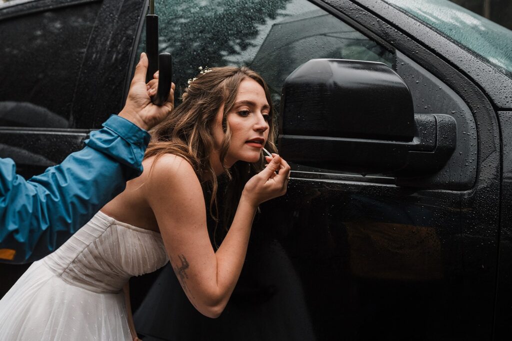 Bride puts on makeup in the car mirror before her forest elopement ceremony