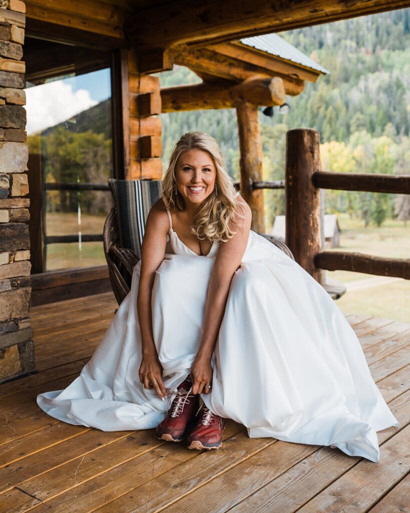 Bride sits on the porch of a Colorado cabin tying her burgundy hiking boots 