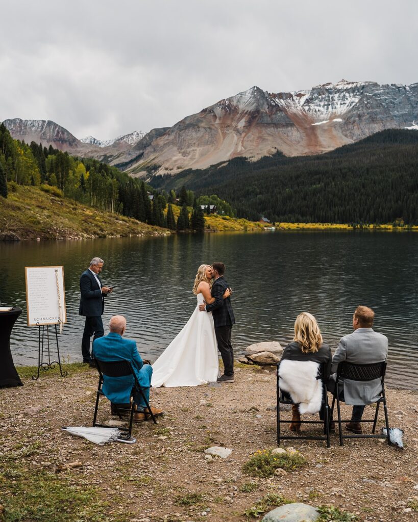 Bride and groom kiss during their elopement ceremony by an alpine lake while guests look on from folding chairs