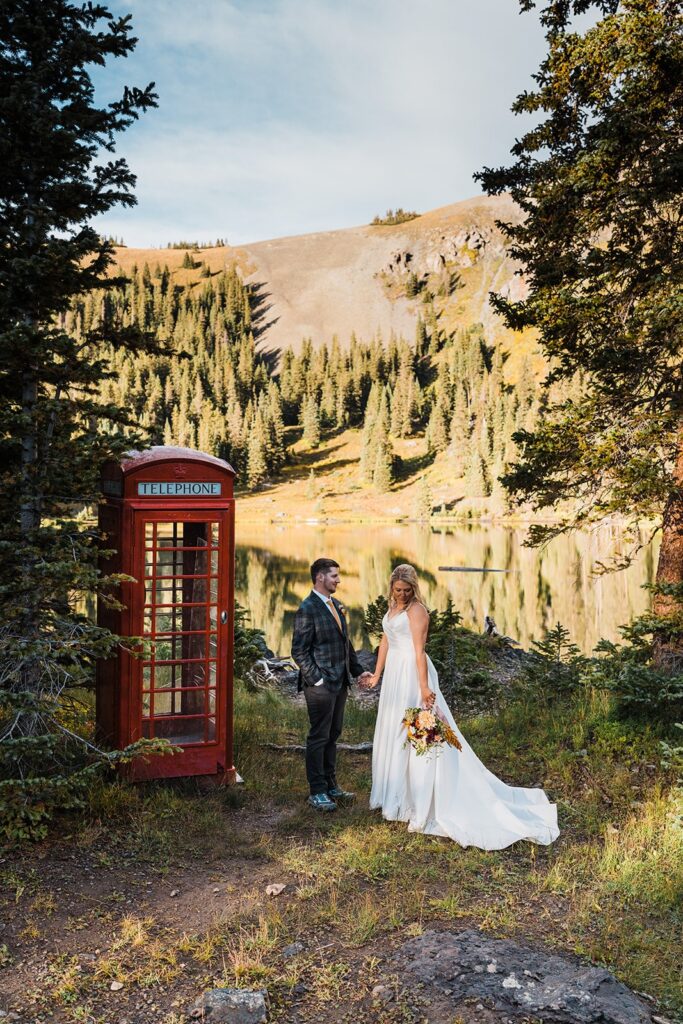 Bride and groom hold hands by a red telephone booth at an alpine lake in Telluride