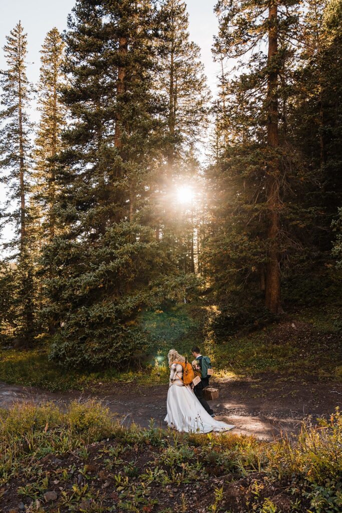 Bride and groom hike down the mountain in their wedding attire while wearing blue and orange hiking backpacks