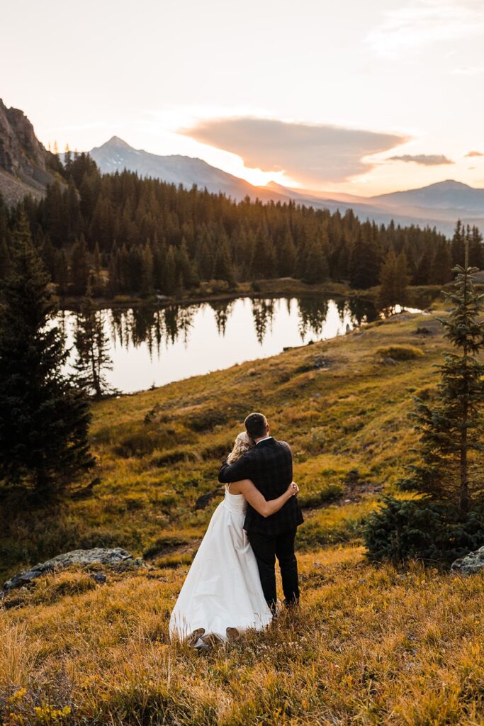 Bride and groom stand on a mountain trail looking out at the sun set over the mountains in Colorado