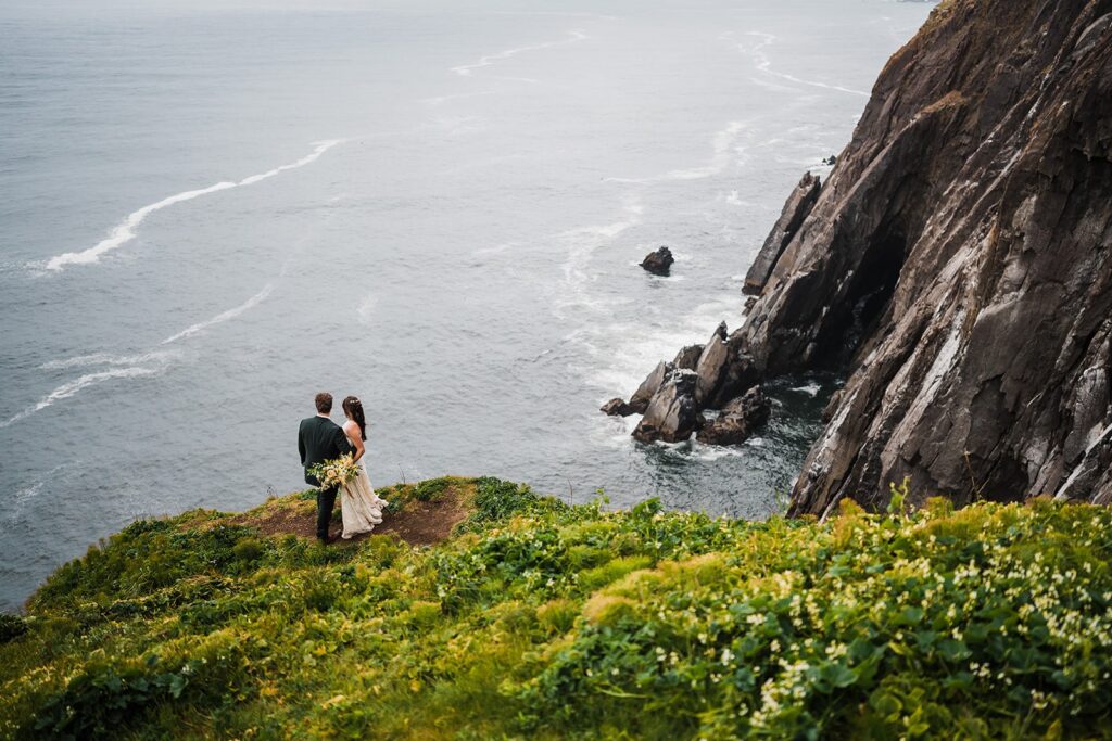 Bride and groom stand on an oceanside cliff looking out at the water during their elopement in Oregon