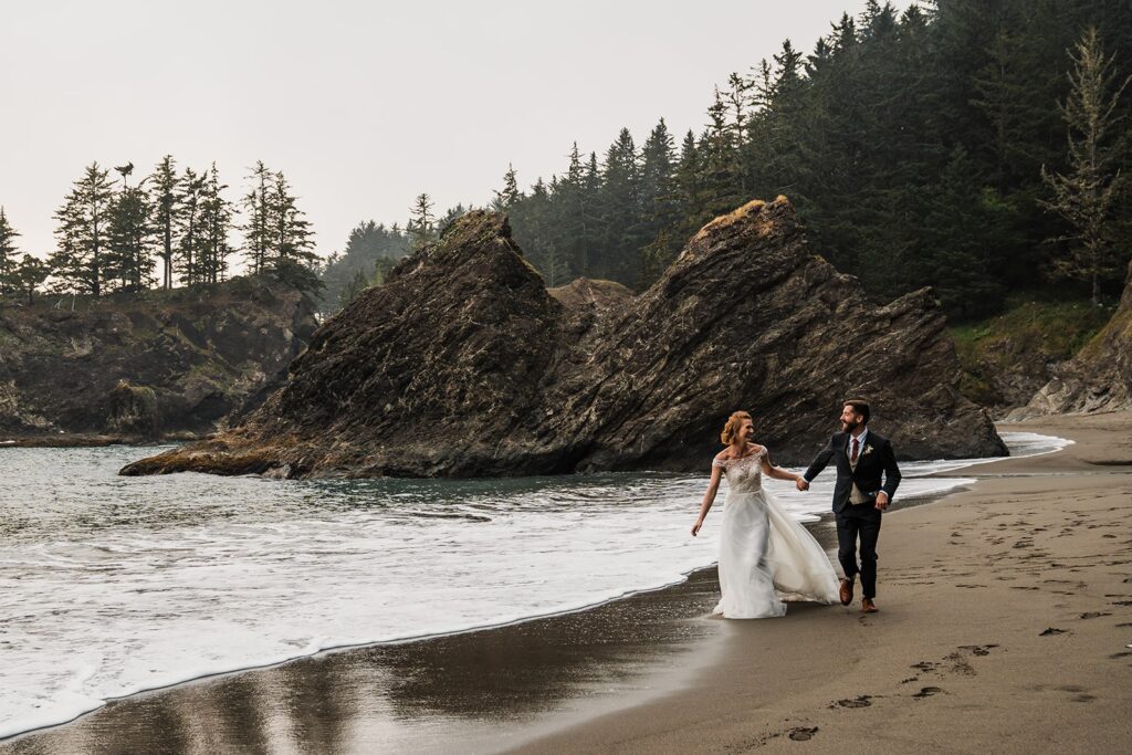 Bride and groom hold hands while running across the beach at their Oregon elopement