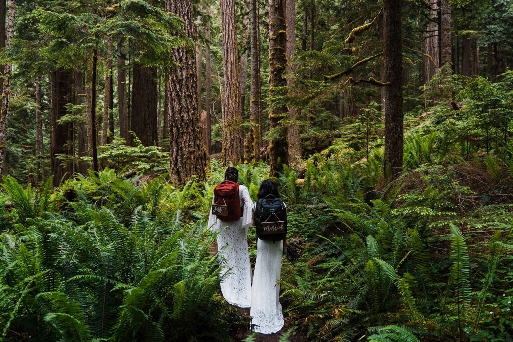 Brides wear hiking backpacks while walking through a forest trail during their elopement in Olympic National Park