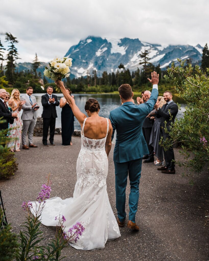 Bride and groom hold hands and wave to their loved ones as they exit their outdoor wedding ceremony in the North Cascades
