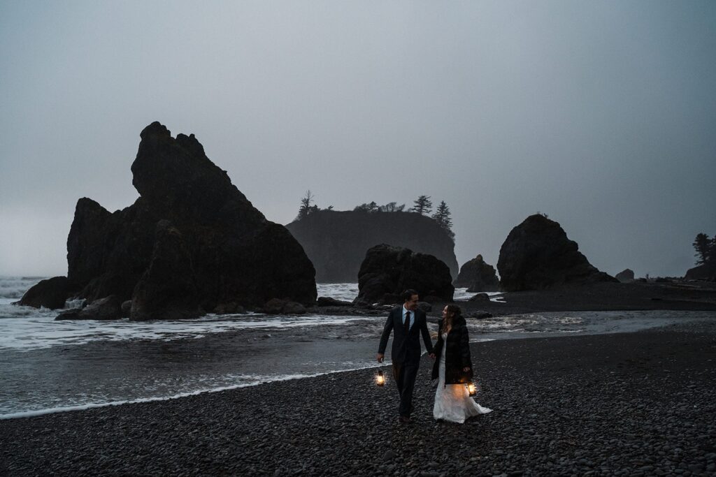 Bride and groom hold lanterns while walking across a rocky beach in Olympic National Park after sunset