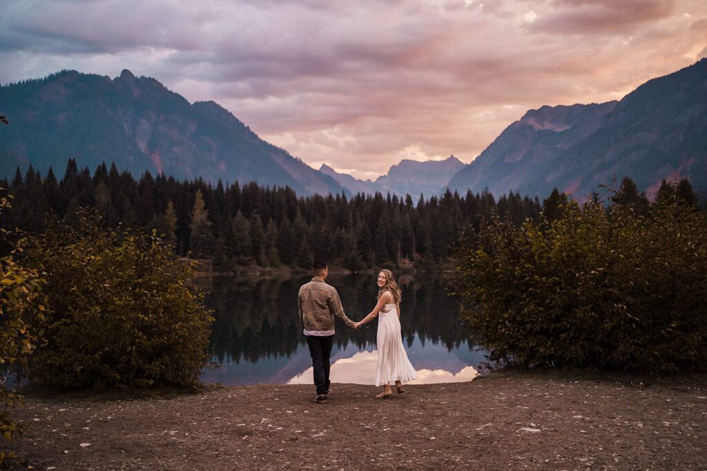 Bride and groom hold hands by a scenic lake during their sunrise adventure session in the PNW
