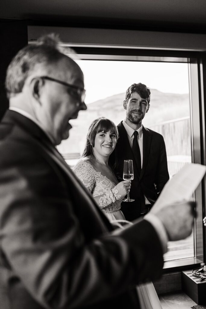 Bride and groom smile while a family member reads a toast at their celebration meal at Hotel Geysir