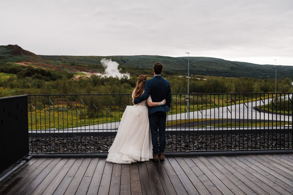 Bride and groom stand side by side looking out at a geyser in Iceland