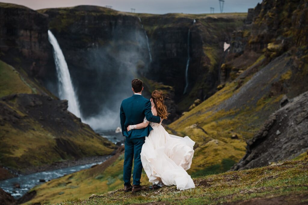 Bride and groom hug while looking out at a waterfall in Iceland