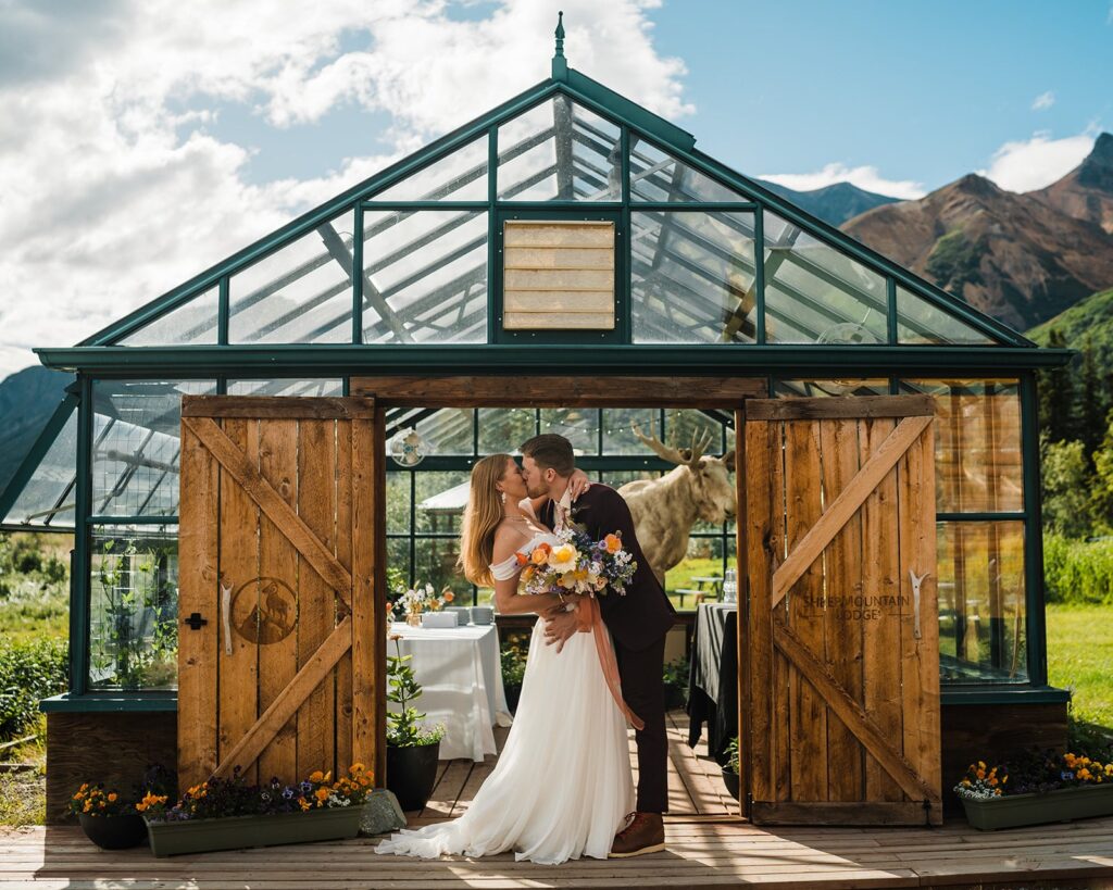 Bride and groom kiss in front of a glass greenhouse as their Sheep Mountain Lodge micro wedding