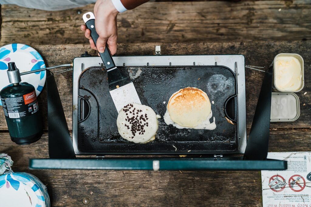 Bride and groom flip chocolate chip pancakes on a camp stove in the forest