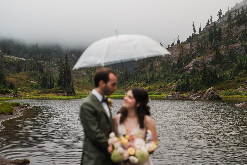 Bride and groom stand under a clear umbrella by Barclay Lake during their rainy wedding 