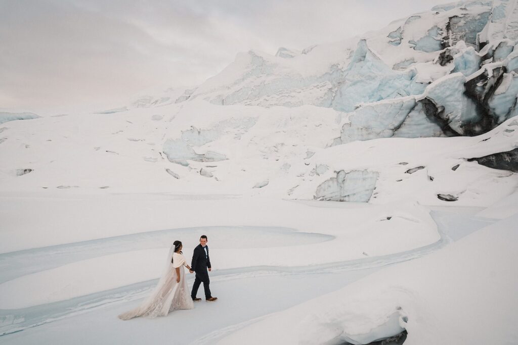 Bride and groom hold hands as they walk across an ice glacier in Alaska