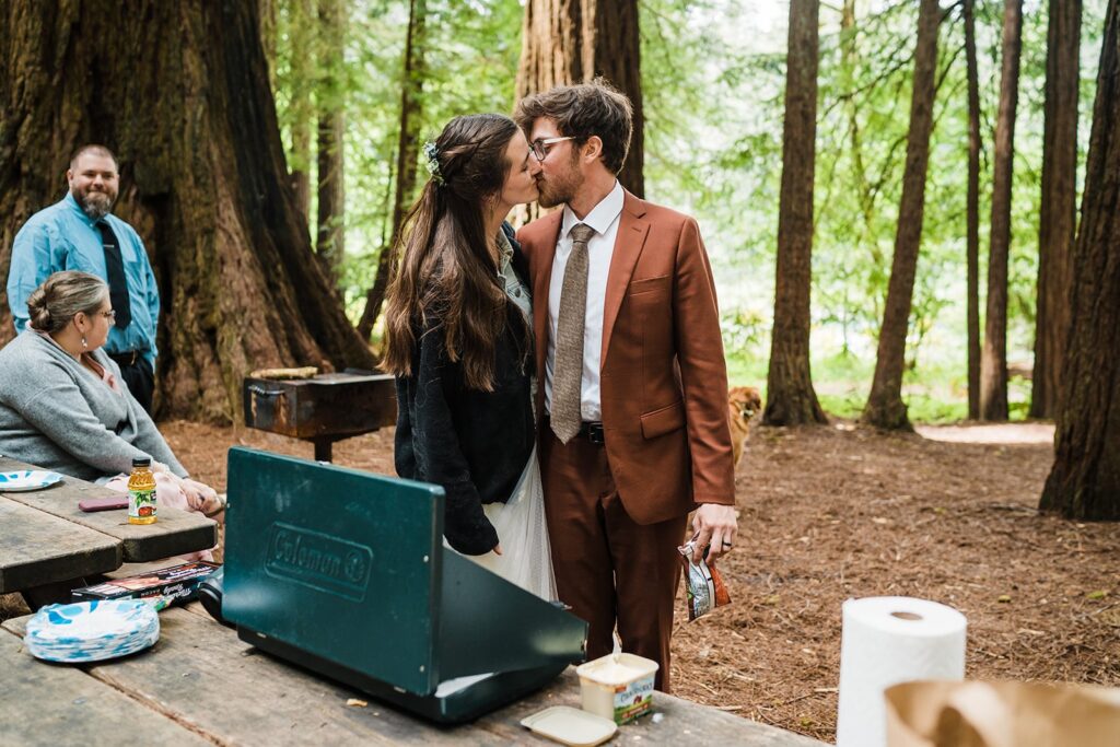 Bride and groom kiss while cooking pancakes over a camp stove at their micro wedding venue at a picnic area