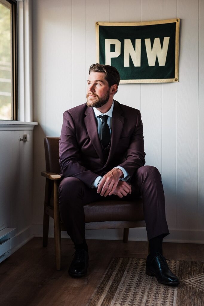 Groom sits on a leather chair in front of a PNW banner after getting ready for his cabin wedding in Washington