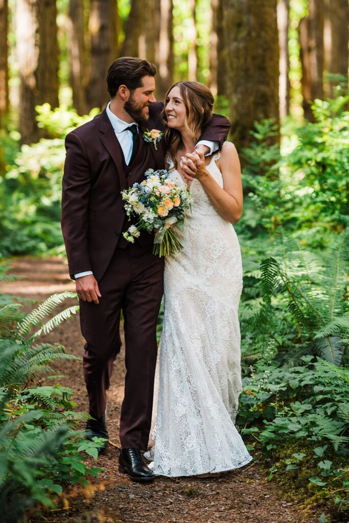 Bride and groom walk through a forest trail with arms around each other 