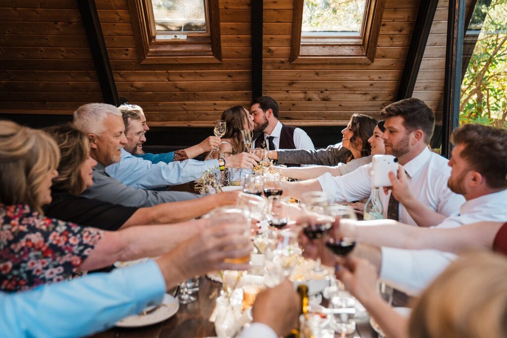 Bride and groom kiss while guests toast at their intimate cabin wedding meal 