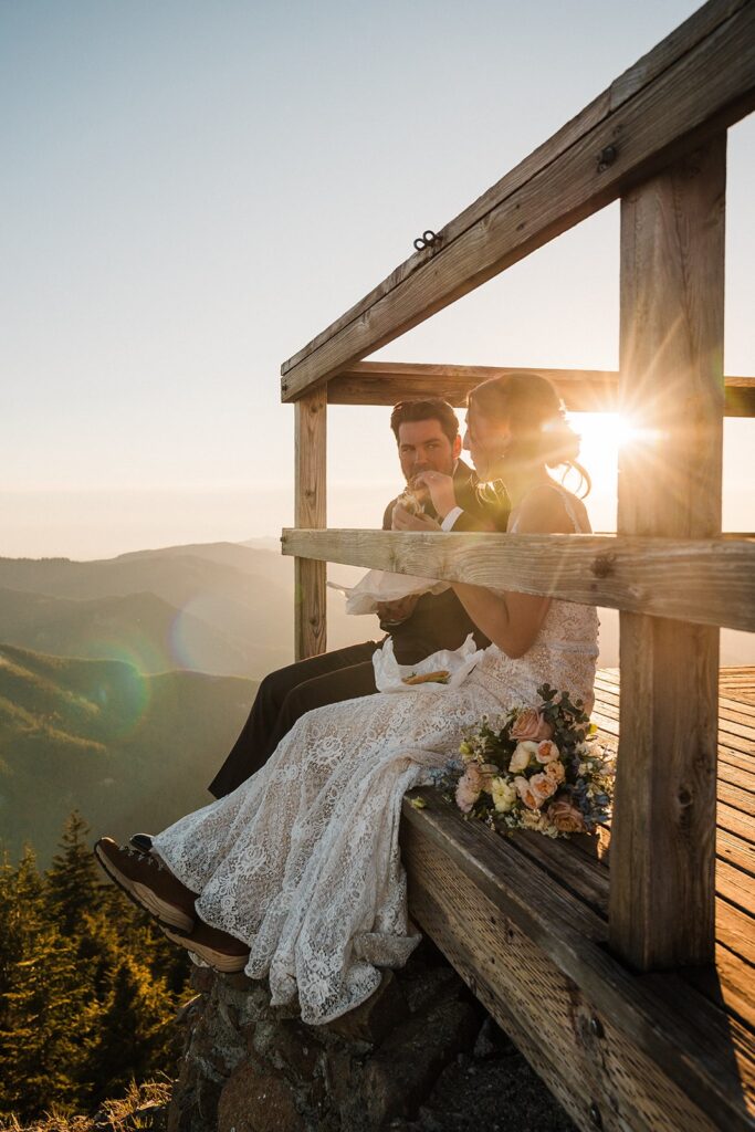 Bride and groom eat pastries while sitting on the edge of a fire lookout near Mount Rainier National Park