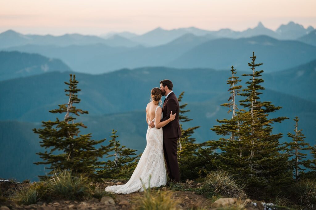 Bride and groom hug on a mountain trail overlooking the mountains surrounding Mount Rainier National Park