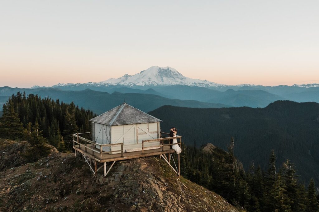 Bride and groom kiss on a fire lookout ledge with Mount Rainier rising in the background