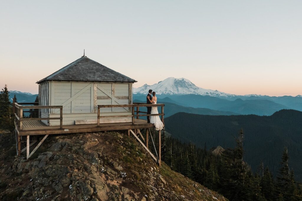 Bride and groom kiss on a fire lookout ledge with Mount Rainier rising in the background