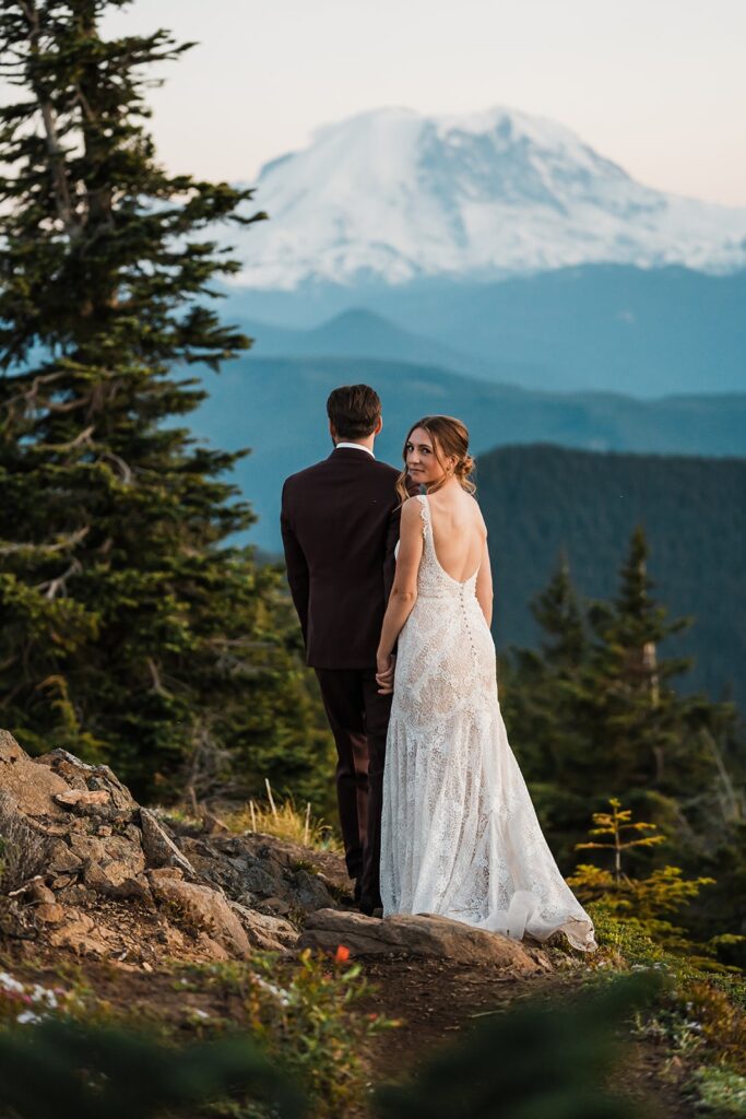 Bride and groom hold hands while walking across a mountain trail in front of Mount Rainier 