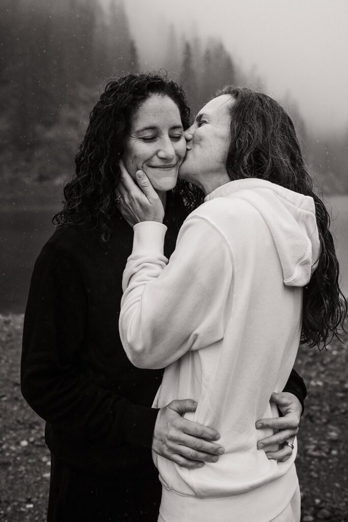 Woman kisses her partner on the cheek in the rain during their LGBTQ elopement sunrise session